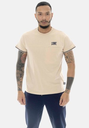 Uomo con tatuaggi su entrambe le braccia, indossa una maglietta beige Leone 1947 e pantaloni scuri, in piedi davanti a uno sfondo chiaro uniforme.