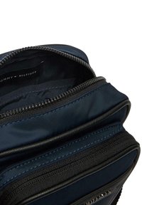 Námořnicky modrá nylonová taška s hladkou texturou, vybavená zipovým uzávěrem a černými doplňky. Vnitřek obsahuje několik přihrádek pro lepší organizaci.