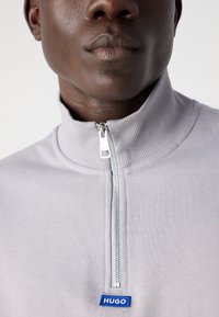 Šedá mikina na zip s vysokým límcem, která má stříbrný zip a modrou nášivku "HUGO" na hrudi. Hladká textura a přiléhavý střih.