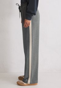 Persoon in grijze wijde broek met een witte verticale zijstreep, zwarte sweatshirt en beige casual schoenen, staand tegen een witte muur.