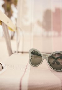 Occhiali da sole a forma di cuore con montature verdi e lenti scure poggiati su una superficie chiara e testurizzata con un motivo a strisce rosa e bianche.