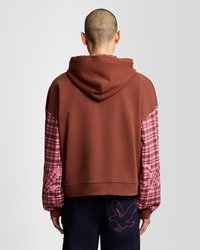 Persona que lleva un sudadera con capucha marrón, con mangas de cuadros rosas y bordados de pájaros en las mangas y en el bolsillo trasero, mostrada desde atrás.