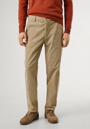 Pantalones de pana beige con corte recto, que presentan líneas texturizadas, bolsillos laterales y cierre de botón. Combinados con zapatillas marrones.