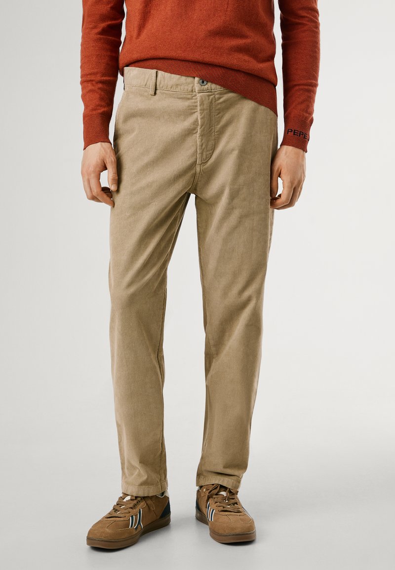 Beige corduroy broek met een rechte snit, voorzien van gestructureerde lijnen, zijzakken en een knoopsluiting. Gemaakt in combinatie met een paar bruine sneakers.