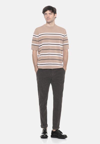 Uomo in piedi con le mani nelle tasche, indossa un maglione corto a righe beige, pantaloni scuri e scarpe eleganti nere, su sfondo neutro.