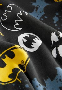 Svart tyg med olika Batman-logotyper i gult, vitt och grått, med en slät textur och spridda blåa färgliknande accenter.