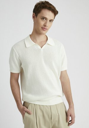 Polo shirt blanc à manches courtes avec un motif tricot texturé et un col contrastant. Porté avec un pantalon ample de couleur claire.
