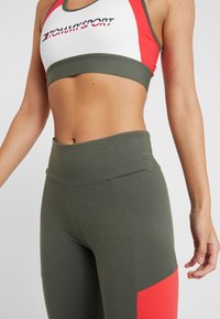 Grüner Sport-BH mit roten und weißen Akzenten, versehen mit einem Logo. Kombiniert mit olivgrünen, hochgeschnittenen Leggings mit einem roten Panel-Detail.