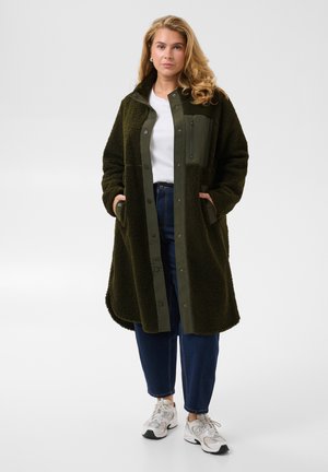 Femme debout portant un long manteau en polaire vert foncé, un t-shirt blanc, un pantalon bleu et des baskets blanches sur un fond clair uni.