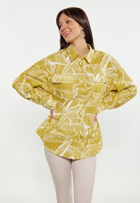 Camicia gialla a maniche lunghe con un motivo a foglie bianche, dal taglio comodo e con due tasche sul petto. Abbinata a pantaloni beige chiaro.