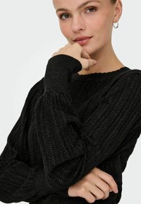 Pull en tricot noir avec texture côtelée, décolleté ondulé et manches longues. Le modèle pose avec la main sur le menton, mettant en valeur le design du pull.