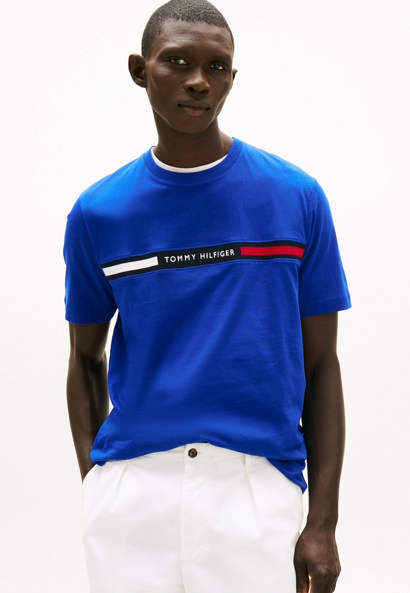 Blå bomulds T-shirt med korte ærmer. Har et horisontalt stribet mønster i hvid, sort og rød med "TOMMY HILFIGER" logoet centreret.