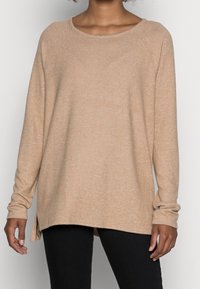 Donna che indossa un maglione lungo beige, ampio e con spacchi laterali, abbinato a pantaloni neri, su uno sfondo bianco uniforme.