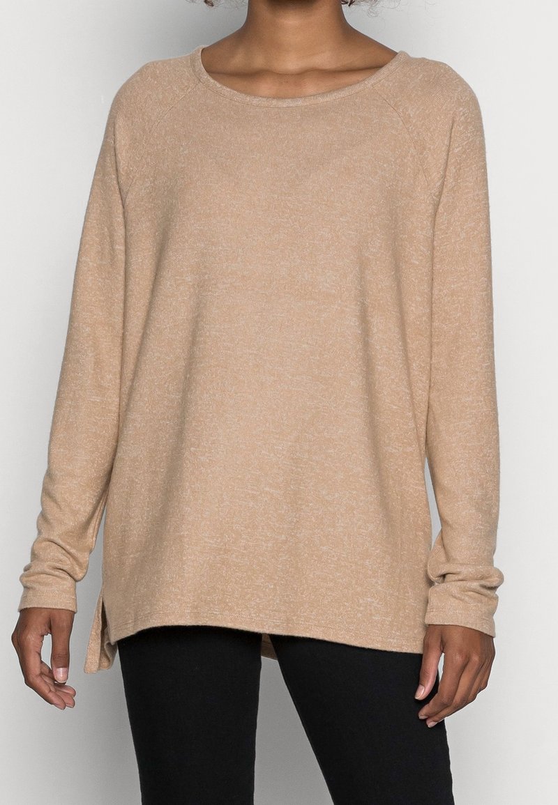 Donna che indossa un maglione lungo beige, ampio e con spacchi laterali, abbinato a pantaloni neri, su uno sfondo bianco uniforme.