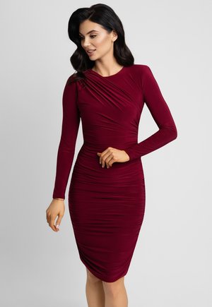 Kobieta w dopasowanej, długorękawowej sukience w kolorze bordo z marszczeniami, pozująca z jedną ręką przy talii, patrząca lekko w dół w lewo.