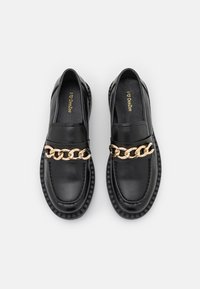 Mocassins noirs avec semelles épaisses et décoration en chaîne dorée sur le dessus, présentés côte à côte sur un fond blanc uni.