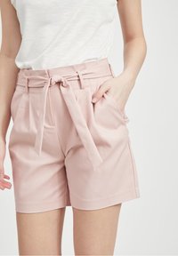 Woman wearing light pink high-waisted shorts with a tied belt and white short-sleeve top, standing with one hand in pocket.