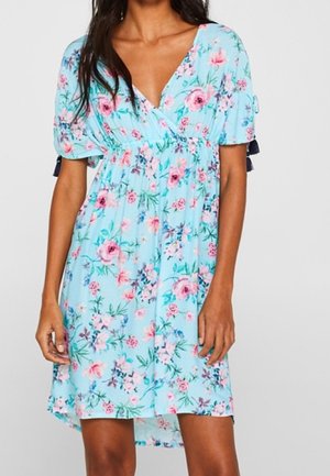 Femme portant une robe fleurie bleu clair avec de courtes manches bouffantes et un décolleté en V, debout, les bras détendus le long du corps.