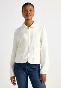 Giacca bianca texturizzata con colletto, chiusura frontale con zip e due tasche a toppa, abbinata a jeans blu. Tessuto morbido con dettagli sottili.