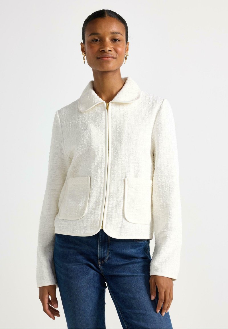 Giacca bianca texturizzata con colletto, chiusura frontale con zip e due tasche a toppa, abbinata a jeans blu. Tessuto morbido con dettagli sottili.