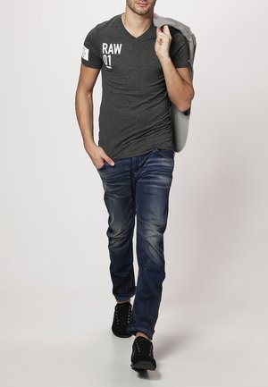 Grå V-ringad T-shirt med vit "RAW 01" tryck, kombinerad med blå jeans och svarta sneakers. Modellen håller en grå jacka över axeln.