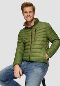 Grüne Pufferjacke mit Reißverschluss, horizontalem Steppmuster und roten Akzenten. Kombiniert mit blauen Denim-Jeans. Weicher Stoff.