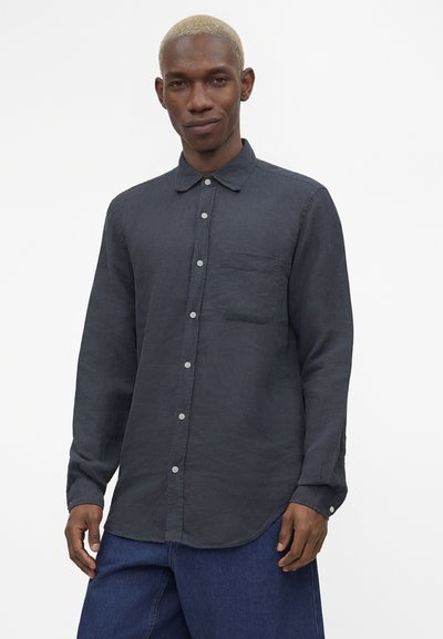 Άνδρας με κοντά ξανθά μαλλιά, φορώντας ένα μακρυμάνικο σκουρόγριμο πουκάμισο με κουμπιά και μπλε παντελόνι, σε απλό λευκό φόντο.