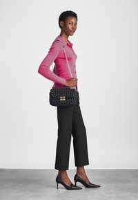 Borsa nera intrecciata con una chiusura in tonalità oro, abbinata a una camicetta rosa con bottoni e pantaloni neri sopra la caviglia, su uno sfondo neutro.