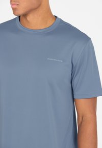 Vaalea sininen urheilupaita, lyhyet hihat, pyöreä kaula-aukko, kosteutta siirtävä kangas, jossa on "ENDURANCE"-logo hillityssä harmaassa painatuksessa.