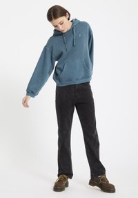 Blauwe katoenen hoodie met een voorzak, losse pasvorm en verstelbare koordjes, gecombineerd met zwarte rechte jeans en bruine schoenen.
