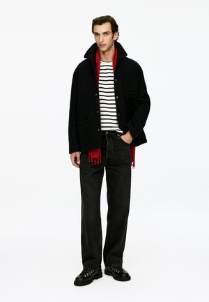 Schwarzer Wollmantel mit rotem Kragen, gestreiftes Hemd darunter, schwarze Jeans und schwarze Schnürstiefel, vor einem weißen Hintergrund präsentiert.