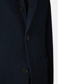 Mörkblå herringbone-blazer med svarta knappar, slagkrage och ett bröstfack. Har texturerad tyg och en strukturerad passform.