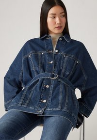 Jachetă din denim cu talie cu curea, buzunare mari și guler clasic. Materialul este de culoare albastru închis cu un finisaj texturat și accesorii din metal argintiu.