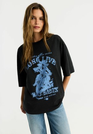Kobieta o długich brązowych włosach, ubrana w luźny czarny T-shirt z niebieską grafiką motocykla i napisem, zestawiony z jasnoniebieskimi dżinsami.