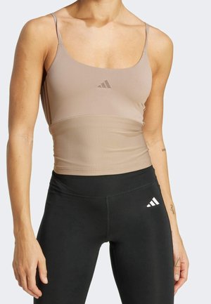 Kobieta w beżowym dopasowanym topie na ramiączkach i czarnych legginsach z wysokim stanem, oba z logo Adidas, stoi na jednolitym tle.