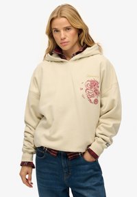 Jonge vrouw met blond haar, die een beige hoodie draagt met rode draakborduursels, gecombineerd met een rood geruit overhemd en blauwe spijkerbroek.
