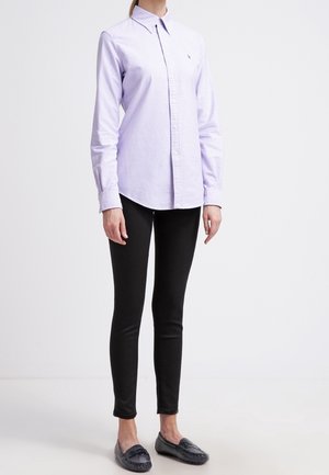 Vrouw draagt een lichtpaarse blouse met knopen, zwarte skinny broek en donkere loafers, staand tegen een effen witte achtergrond.