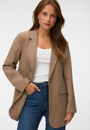 Femme aux longs cheveux bruns porte un blazer beige, un haut blanc, un jean bleu et des boucles d'oreilles en perles, debout avec une main dans la poche.