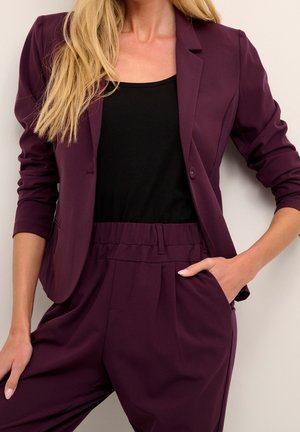 Mujer con blazer de color púrpura oscuro y pantalones de talle alto a juego, combinados con una blusa negra, con una mano en el bolsillo, posando contra una pared clara.