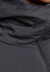 Material einer dunkelgrauen Daunenjacke in Nahaufnahme, mit hohem Kragen, glatter Textur und sichtbarem Reißverschlussdetail an der Seite.