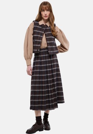 Femme portant un chemisier beige à manches bouffantes, un gilet marron à carreaux, une jupe plissée assortie à carreaux, des chaussettes foncées et des chaussures en cuir marron.