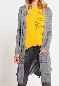 Grå lång cardigan med öppen front och två frampocket över en ljusgul t-shirt med svart text och ett hjärtformat mönster.