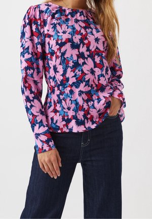 Vrouw draagt een roze bloes met lange mouwen en bloemenmotief met blauwe en rode accenten, gecombineerd met donkerblauwe spijkerbroek, staand met één hand in de zak.