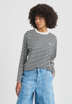 Vrouw met krullend haar, gekleed in een zwart-wit gestreept shirt met lange mouwen en blauwgepatenteerde wijde jeans, staand met één hand in de zak.