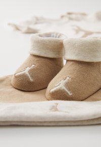 Botitas de bebé de algodón en tono marrón claro con un puño acanalado, que presentan un logo Jumpman bordado en blanco. Diseño sencillo sobre un fondo de tela beige.