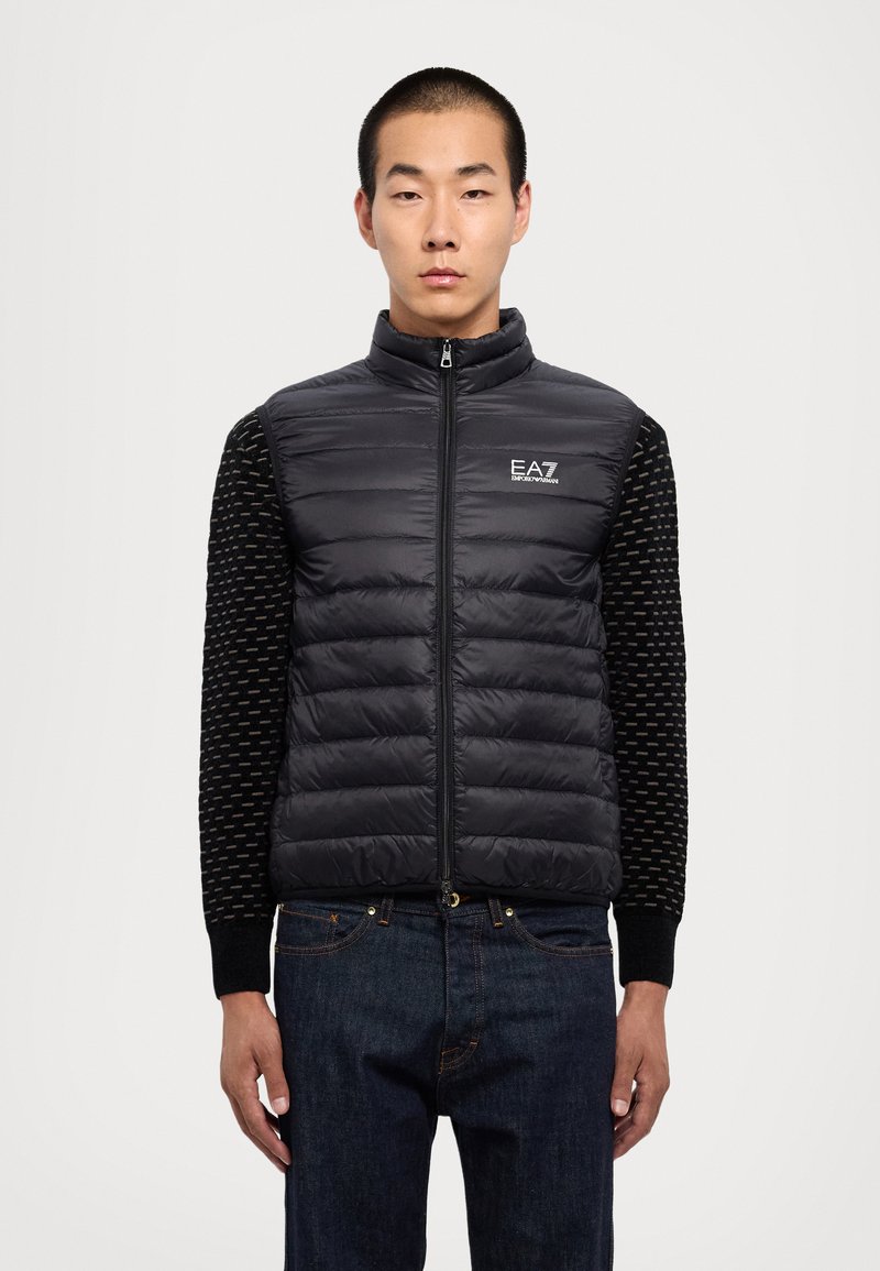 Gilet rembourré noir avec un col haut, une fermeture éclair à l'avant et des manches tricotées noires à motif texturé. Il présente un petit logo EA7 sur la poitrine.
