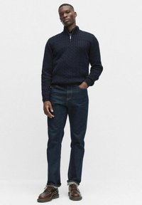 Marinblå kabelstickad zip-up tröja, mörka jeans och bruna snörskor. Tröjan har en hög krage och strukturerad mönster.