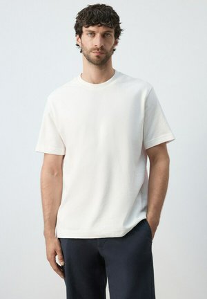 Hombre con cabello rizado y oscuro, vistiendo una camisa blanca lisa de manga corta y pantalones oscuros, de pie frente a un fondo gris claro.