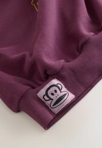 Sweatshirt violet cu o textură moale, având manșete cu nervuri și o aplicație pătrată proeminentă pe care este reprezentat un logo de maimuță în negru pe un fundal roz.