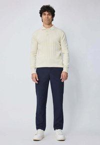 Homme debout portant un pull polo à manches longues côtelé crème, un pantalon bleu marine foncé et des baskets blanches sur un fond clair uni.